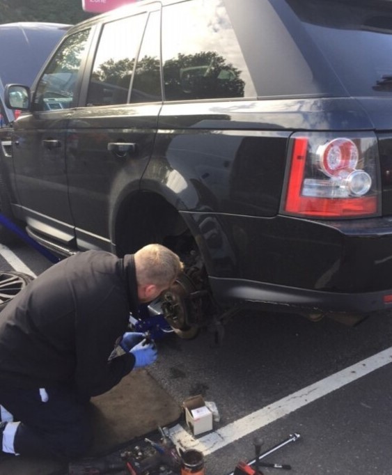A man repairing a car in a parking lot. He is focused on fixing the brakes of the vehicle.